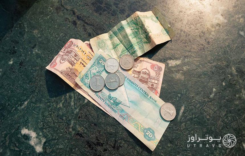 سه اسکناس درهم امارات روی پس‌زمینه‌ای تیره‌رنگ و چند سکه روی آن‌ها